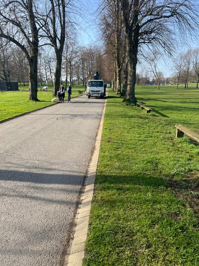 The image depicts a park scene in Lurgan, United Kingdom, on a clear, sunny day. A paved, grey asphalt path, bordered by a light-colored curb on its right, extends into the mid-ground, flanked by an avenue of mature, bare deciduous trees. Green grass covers the ground on both sides of the path. On the right side of the path, several wooden park benches are visible on the grass.
In the mid-ground, a white utility truck, bearing the license plate VGN WCJ, is positioned on the path facing away from the viewer. An individual, dressed in dark clothing, stands on equipment mounted on the rear of the truck. To the left of the path, two individuals are standing: one woman with light hair, wearing a dark jacket over a white top and dark trousers, holds a leash attached to a small, light-colored dog. She is positioned near a man wearing a blue jacket and dark trousers. In the background, beyond the trees, further open grassy areas and what appear to be recreational structures are discernible. Shadows from the trees are cast across the path and grass, indicating a low sun angle.