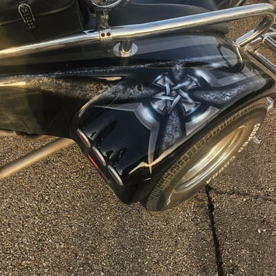 A close-up, high-angle shot captures a black motorcycle fender and wheel. The fender features an airbrushed design of a silver, multi-pointed star or emblem with shaded metallic effects. The tire is a Cooper Cobra, with white lettering visible on its sidewall. The motorcycle is parked on a textured, gray asphalt surface that is cracked in several places. The background consists of the motorcycle's leather saddlebag, chrome accents, and a reflective metallic bar. No people are present, and the scene appears to be outdoors in daylight. This image was taken in Illescas, Spain.