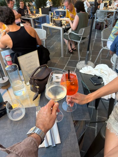 The image depicts an outdoor dining scene at a restaurant in Loveno, Italy. In the foreground, a man's arm and hand, adorned with a silver watch, hold a stemmed glass of white wine. Across the table, another person's arm and hand, wearing a white lace garment on their arm, reach for a stemmed glass containing an orange-hued drink, possibly an Aperol Spritz. Between the two glasses are silverware and napkins. The table is covered with a dark gray cloth, and a menu titled "Food DA CONDIVIDERE TO SHARE" is visible. To the left, a clear glass bottle, likely water or spirits, stands next to a small, clear glass globe. In the mid-ground, other diners are seated at tables, some interacting with food and drinks. The background shows more tables, chairs, and the architecture of the establishment, hinting at a charming Italian setting. The overall ambiance suggests a leisurely meal or drinks outdoors.