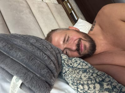 The image depicts an adult male lying shirtless on white bedding in a bedroom setting, presumably in Business Bay, United Arab Emirates. The man is positioned on his side, facing the camera, with his head resting on two distinct pillows: a grey, plush, ribbed pillow/blanket on the left and a smaller decorative pillow with a blue, white, and brown patterned knit cover directly under his head. He has short hair with visible grey streaks, a full beard, and a mustache. He is smiling broadly, with his right eye closed in a wink or squint, and his left eye open, looking directly at the viewer. A tattoo of three vertical East Asian characters is visible on his right shoulder/upper back. The background features a light beige upholstered headboard with vertical panels, a dark brown wall or paneling, and a vertical brass-colored lighting fixture with two white cylindrical bulbs. A white, plain pillow is partially visible to the upper right of the man's head. The grey plush item on the left has a white tag with text and a logo, partially visible.