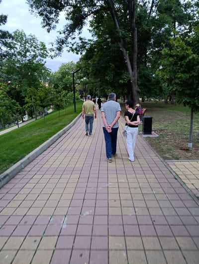 The image depicts a paved pathway in a park with lush green trees and grass. Several people are walking on the pathway. In the foreground, a couple is walking away from the camera, with the man in a blue t-shirt and jeans, and the woman in a black top and white pants. Further down the path, a man in a light green t-shirt and jeans is also walking away. The park appears to be a public space, likely in the city of Kelerlesskaya, Russia, given the context. The lighting suggests it is either early morning or late afternoon.