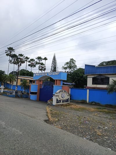 The image depicts the entrance to "Unidad Educativa Eloy Alfaro", a school in Quevedo, Ecuador. The school's name is prominently displayed above the entrance gate in blue lettering. The gate itself is dark blue with decorative metalwork. A sign with the school's name, "Colegio Fiscal Eloy Alfaro", is on the right side of the entrance.

The architecture includes a mix of one and two-story buildings painted in white and blue. A blue wall runs along the perimeter of the property. Palm trees line the front of the school, and several power lines are visible overhead. The street leading to the entrance is paved, with patches of dirt and gravel visible. The sky is overcast, suggesting a cloudy day.