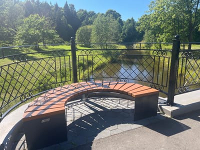A curved, slatted wooden bench with black metal supports sits on a paved area in front of a decorative black metal fence. The fence overlooks a body of water, likely a river or lake, which is bordered by a grassy slope and a dense forest of green trees. The sky is clear and blue, and sunlight casts distinct shadows on the pavement. The scene appears to be in a park or a scenic overlook. This is likely the bench on the bridge over the Pühajõgi river in Toila, Estonia.