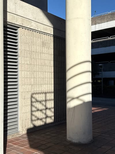 An abstract composition features a cylindrical concrete column horizontally aligned, casting curved shadows upon a textured wall beneath. Below, a horizontal grating with parallel black slats defines a lower plane. A complex geometric shadow resembling a window frame overlaps the textured wall, which transitions into a brick pattern on the right side. The entire scene is bathed in bright light, highlighting the interplay of shadows and textures.