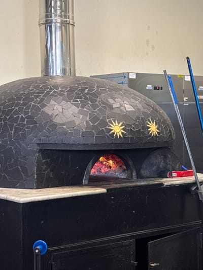 The image shows a close-up of a black outdoor pizza oven with a mosaic-tiled dome. The dome is decorated with two golden sun emblems. A stainless steel chimney extends from the left side of the oven, and a fire burns within the oven's opening, with glowing embers visible. The oven appears to be made of metal with a stone or concrete base. The background shows a pale, textured wall and a portion of what looks like an air conditioning unit with a digital display. There are also some blue poles or handles extending from the top left of the frame. The scene suggests an outdoor cooking environment, possibly a restaurant or a home patio. The lighting appears to be natural, but the time of day is not definitively indicated. There are some smudges and marks on the wall and the oven, suggesting use. No people are visible in the image. Text is visible on the air conditioning unit, but it is too blurry to read. The location context is Carini, Italy.