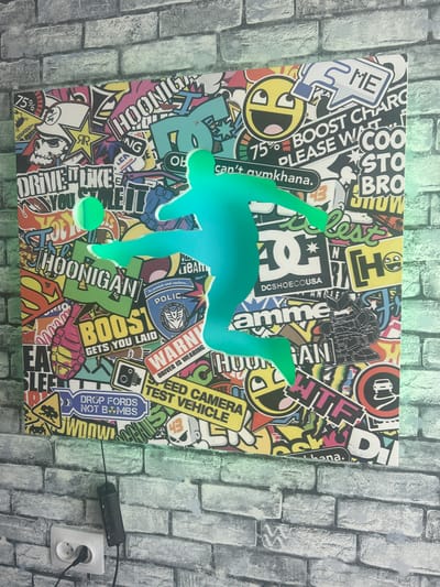 The image shows a vibrant sticker collage on a canvas depicting a silhouette of a soccer player in a green glow. The collage is filled with various stickers featuring logos, text, and cartoonish images. Visible text includes "HOONIGAN," "BOOST CHARGE," "DRIVE LIKE YOU STOLE IT," "POLICE," "WARNING," "SPEED CAMERA TEST VEHICLE," and "DROP FORDS NOT BOMBS." The canvas is mounted on a brick-patterned wall. To the left of the canvas, a white electrical outlet with two plugs is visible, and a black power adapter is attached to the wall below the canvas. The scene is illuminated by a green light, which casts a glow on the soccer player silhouette and the surrounding wall. There are no people visible in the image. The location is not specified within the image itself.