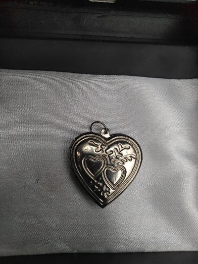 A silver-toned, heart-shaped pendant or locket rests on a light grey, wrinkled satin-like fabric lining inside a black, textured rectangular case. The pendant features an embossed design with two smaller heart shapes near the bottom, flanked by stylized leaf or scroll motifs. A small, dark-colored bail is attached at the top of the heart for suspension. The image depicts this object located in Лебяжье ауылдық округі, Kazakhstan. No individuals, actions, or external environmental details are present.