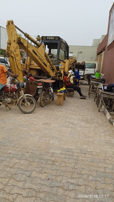 This image depicts an outdoor scene in Ijaiye, Nigeria, on 2026/1/30 at 12:11. The foreground is a paved area with interlocking concrete pavers. A large, yellow Ford 655 backhoe loader, bearing the "FORD" logo on its boom and cab, is stationary on the left side of the frame. Several individuals are present: a man in an orange shirt is partially visible on the far left, near a red motorcycle. Two other motorcycles are parked next to the backhoe; one features a brown sidecar. A woman in a red shirt and black pants sits on a wooden bench on the right, holding a baby wrapped in yellow fabric. Another wooden bench with yellow clothing draped over it is beside her. Two rudimentary wooden tables, laden with various items including a blue bucket, are positioned further right. Multiple men are visible around the backhoe, one in a camouflage-patterned shirt and another in a blue shirt. A young child in a red shirt is partially visible near the backhoe. In the background, a white van with "DURING" and "NG AREA" text is partially visible behind the backhoe, and structures including a reddish-brown building with a partially visible poster are present. The sky is overcast.