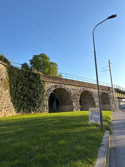 The image displays a picturesque scene featuring a historic stone railway viaduct in Hainburg an der Donau, Austria, under a brilliant clear blue sky. The viaduct is characterized by its robust stonework and several rounded arches, some of which are partially adorned with dense, dark green ivy.
A expansive, well-kept lawn with bright green grass occupies the foreground and extends along the base of the viaduct, displaying distinct shadows that suggest the time of day is either mid-morning or late afternoon. To the left, a tall, modern streetlamp with a curved arm reaches into the sky. Multiple thin overhead lines, indicative of an electrified railway system, crisscross the upper portion of the frame. The paved surface of a road or path can be seen running along the top of the viaduct, with glimpses of other structures or vehicles in the distance.
In the grassy area to the right, a white sign on a pole is prominently displayed. The text on the sign advertises "DD's MAIBAUMFEST" (Maypole Festival) scheduled for "30.04.2023." Further details on the sign highlight "EINTRITT FREI!" (Free Entry!), the name "BUTTLER," and offerings of "BIER ODER SEKT" (Beer or Sparkling Wine), "MUSIK" (Music), and "SPEISEN & GETRÄNKE" (Food & Drinks). It also mentions "Für gute Stimmung sorgt..." (For a good atmosphere provides...) followed by a partially visible band name. No individuals are present in the immediate scene, but the sign indicates an upcoming community event. The weather is sunny and clear, contributing to a serene atmosphere.