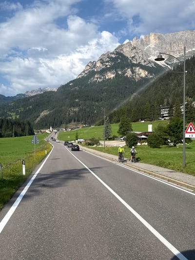 The image depicts a scenic mountain road in Mazzin, Italy, under a partly cloudy sky. A two-lane asphalt road with white lane markings curves through a lush green valley. On the right side of the road, two individuals are cycling on a paved path adjacent to the road. One cyclist wears a bright yellow shirt, and the other wears a dark shirt. Further down the road, several cars are visible, navigating the gentle curves.
The landscape is characterized by steep, forested mountains that rise dramatically in the background. The trees appear dense and dark green, creating a stark contrast with the lighter green of the meadows and fields that flank the road. In the distance, a cluster of buildings, likely part of a village, is nestled in the valley. A prominent church steeple with a white spire is visible among the buildings. A tall, modern street lamp with a curved arm extends over the road on the right side.
To the far right, a red and white triangular traffic sign indicates a sharp curve ahead, with the notation "x 2" suggesting a double curve or increased hazard. The overall atmosphere is one of a serene, picturesque mountain environment, with recreational activity taking place alongside vehicular traffic.