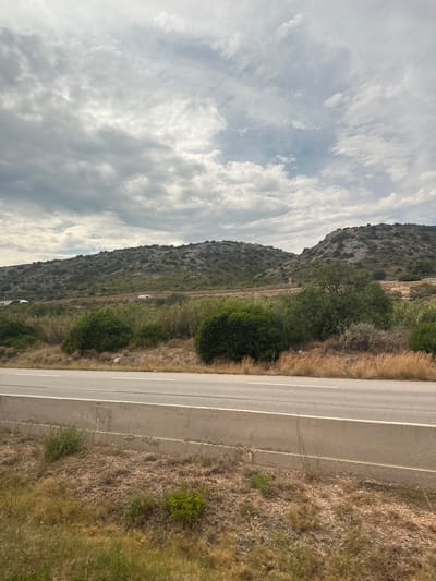 This media file shows a view of a road and a hillside, likely taken from a vehicle traveling along the road. The scene is outdoors and appears to be daytime, with a cloudy sky. The road is paved and has white lane markings. To the left of the road is a grassy and rocky hillside covered in sparse vegetation. There are a few white vehicles visible on the side of the road. In the distance, there are signs on poles along the hillside, one of which appears to be a speed limit sign with "50" and "60" indicated. The overall setting is rural and mountainous.