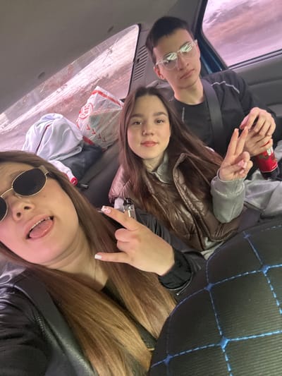 The image shows three young adults posing for a selfie inside a vehicle. The person in the lower portion of the frame is wearing round sunglasses and sticking their tongue out. Above them is another person pointing down with their index finger. The third person, situated towards the top left of the frame, is wearing glasses and looking towards the camera while holding a beverage. Other details include clothing items, a plastic bag with Russian text, and a partial view of the vehicle's interior. The location within the city of Ledukhi, Russia cannot be verified from the image alone.