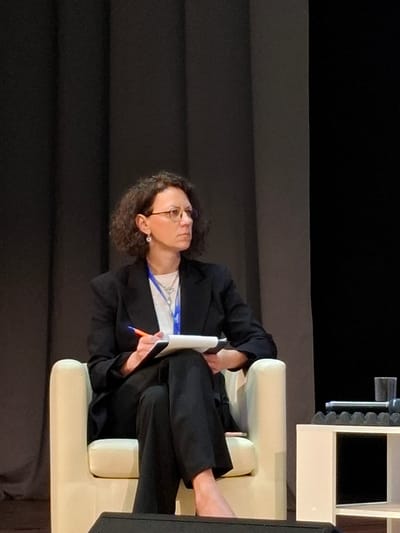 A woman with curly brown hair and glasses sits in a cream-colored armchair on a stage. She is wearing a black suit jacket over a white shirt and black pants. She holds a white notepad and an orange pen in her hands, appearing to be taking notes or writing. She is looking to her right, off-camera. The background is a dark gray curtain. To the right of the armchair is a white table with a glass of water and some electronic equipment on it. The bottom of the image shows a black speaker. The image does not provide any specific information about the location within the city of Kungur, Russia.
