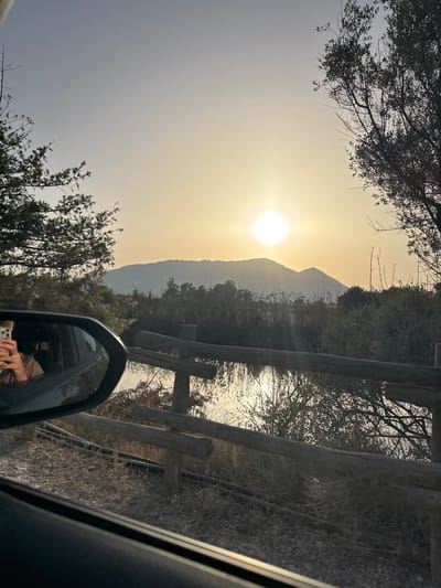 A photograph taken from inside a vehicle shows a person's reflection in the side-view mirror, holding a mobile phone. The background features a serene landscape with mountains silhouetted against a bright, hazy sunset. A rustic wooden fence runs horizontally across the mid-ground, separating a dry, grassy area from a body of water reflecting the sunlight. Trees frame the scene on the left and right, with sparse foliage contributing to the autumnal or dry season atmosphere. The overall impression is one of a tranquil, natural setting at dusk, likely near Orosei, Italy, given the mountainous terrain and Mediterranean vegetation.