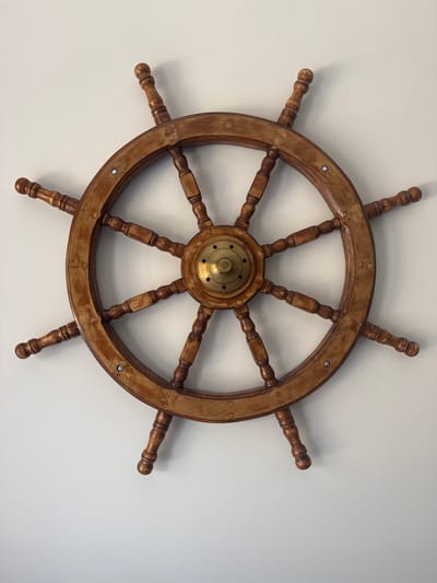 This media file shows a wooden ship's wheel against a plain white background. The ship's wheel is made of a light brown wood and has a brass centerpiece. It has a large outer rim, with eight spokes connecting to the rim from the central hub. Each spoke has a decorative wooden bead design. Extending from the outer rim are eight handles, also made of the same wood, which are cylindrical in shape and rounded at the ends. These handles are positioned around the wheel, spaced evenly apart. The brass centerpiece at the hub of the wheel has a circular design with several small holes around its perimeter and a central bolt or screw. There are also several visible screws or rivets attaching the spokes to the outer rim. The lighting suggests a well-lit indoor environment, possibly with overhead lighting, as there are no strong directional shadows, and the white background appears evenly illuminated. The image is taken from a top-down perspective, looking directly at the ship's wheel. There is no visible text in the image. The scene is simple and focuses solely on the ship's wheel as an object.