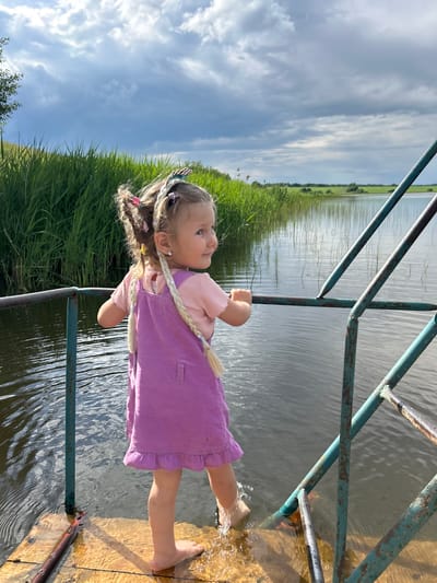 A young girl with blonde hair styled in braids, wearing a pink t-shirt and a purple pinafore dress, stands on a wooden platform next to a body of water. She is reaching her foot into the water, creating a splash. The girl is looking up and to her left with a curious expression. The background shows a vast expanse of water, reeds or tall grass along the bank, and a cloudy blue sky. A rusty metal railing separates the platform from the water. The scene appears to be outdoors during the daytime, with good lighting suggesting it could be late afternoon. The overall atmosphere is one of childhood exploration and enjoyment of nature in a rural or semi-rural setting, possibly near a lake or river in Верх-Рождество, Russia.