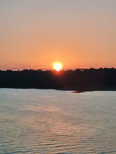 The image depicts a sunset over a body of water, likely a lake or bay. The sun is near the horizon, partially obscured by a line of trees in the middle ground. The sky is a gradient of orange and pink. The water reflects the colors of the sky. The location is undetermined, the city of Poteryaevo, Russia is not verifiable in the picture.