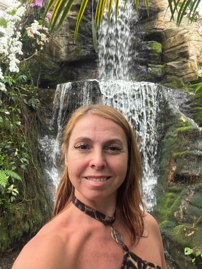 A woman with auburn hair and fair skin takes a selfie in front of a waterfall in what appears to be a botanical garden. She is smiling and her shoulders and upper chest are visible. She is wearing a leopard-print top and a silver pendant necklace with a star symbol. The waterfall is multi-tiered and surrounded by lush greenery, moss-covered rocks, and orchids. The location is identified as Pong Yaeng, Thailand.