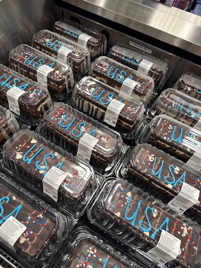 The image shows a close-up, overhead view of several individually packaged chocolate mousse cakes on display, likely in a retail environment such as a grocery store or bakery. The cakes are rectangular and are decorated with blue icing that spells out "USA" on each. They are also adorned with red, white, and blue sprinkles, and swirled chocolate and white icing. The cakes are housed in clear plastic containers with white labels indicating nutritional information and product details. The background is dominated by the reflective surface of a stainless steel display case and hints of other products on shelves above. The setting appears to be the City of Yonkers, United States of America, suggesting a patriotic or holiday-themed display. There are no people visible in the image.