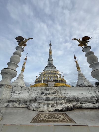 The image shows a view from a low angle, looking up at a white, multi-tiered stupa with golden decorative elements. Two ornate, gray, layered pillars topped with golden mythical bird sculptures flank the stupa. Smaller, similar white stupas with gold accents are visible on either side of the main structure. The stupa is adorned with golden trim and yellow fabric draped around its lower levels. The ground in front of the stupa is paved with light-colored tiles, and a patterned rug is placed on the tiles. The sky above is overcast with gray clouds. This scene is located at Wat Phra That Mae Yan in Ban Sadet, Thailand. No people are visible in the image.