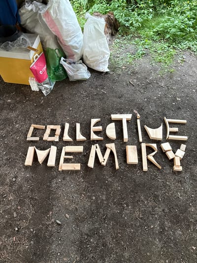 In Cheposh, Russia, on a dirt ground surface covered with pine needles and small stones, wooden letters are arranged to spell "COLLECTIVE MEMORY." To the left of the letters, a yellow cooler is visible, with several large white and pink sacks behind it, along with green and pink shopping bags and a black pot. The background features lush green foliage and a tree stump, indicating an outdoor, possibly forested, environment.
