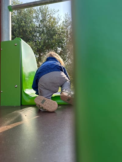 A child wearing a blue jacket and striped pants is positioned on a green slide at a playground. The child's feet, clad in tan-colored shoes with green accents, are visible on a gray surface adjacent to the slide. A portion of a green structure obscures the view above the child. Trees are visible in the background. This image appears to have been taken in Longone al Segrino, Italy.