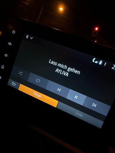 The image captures a close-up, diagonally oriented view of an active automotive infotainment system screen at night, set within the city of Kassel, Germany. The screen displays a music player interface featuring the song "Lass mich gehen" by the artist "AYLIVA." Key elements on the screen include playback controls: a highlighted orange "Player" button, a "Liste" (List) button, and icons for shuffle, repeat, rewind, pause, and fast forward. Along the left edge are touch controls for power, volume up, volume down, and home. The top status bar indicates Bluetooth connectivity, a 'V' symbol, cellular signal strength, battery level, and numerical data "1 2:". The immediate foreground and background are dark, with ambient light sources visible reflecting on the screen and from outside the vehicle. A prominent red rectangular light, resembling a traffic light, is visible in the upper right background, while a softer, circular orange light appears in the upper left. No people are visible.