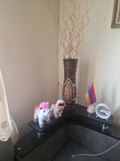 The image displays a vignette of a room, likely a living space, in Nor Kharberd, Armenia. The scene is dominated by a tall, dark, ribbed vase filled with thin, dried branches that curl outwards. The vase itself is decorated with a central oval inlay featuring intricate patterns, possibly of metal or polished wood, with small, reflective embellishments.
To the right of the vase, perched on a dark, modern L-shaped table, are several items. A small Armenian flag, displaying red, blue, and orange stripes, is propped up. Beside it, a clear container holds a few pink, spherical objects, possibly candies or decorative items. Further along the table is a white, crescent-shaped object that appears to be a modern lamp or speaker.
Two plush toys are also visible on the table. One is a white unicorn with a pink mane and tail, and grey hooves. Next to it is a smaller, orange and white robot-like toy with black accents. A white charging cable snakes across the dark table surface, disappearing to the right.
In the upper left corner, a portion of a framed picture is visible, suggesting wall decor. The overall impression is that of a decorated corner of a room, with personal touches like the toys and flag. The lighting suggests it is daytime, with natural light illuminating the scene from an unseen source. The wall behind the objects appears to be a neutral, light color, with some visible cracks.