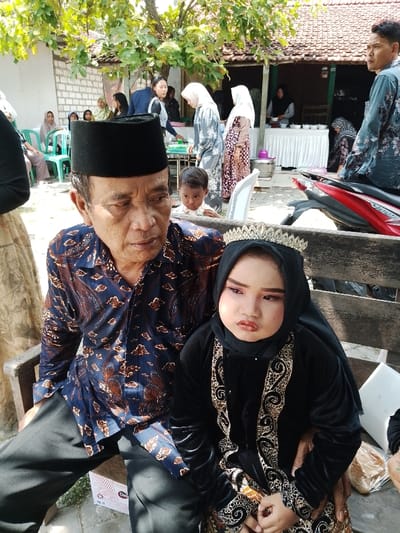 This image depicts an outdoor gathering in Juwana, Indonesia, under a tree canopy with direct sunlight. In the foreground, an older man with a dark complexion, wearing a black songkok and a blue and brown patterned batik shirt over black trousers, is seated on a wooden bench. His gaze is directed forward and slightly downward. His right arm is behind a young child seated next to him, and his right hand rests on the child's left knee. The child, appearing to be a young girl, wears a black hijab, a gold-colored tiara, and a black velvet-like dress with prominent gold embroidered scrollwork. Her face is made up with pink eyeshadow and lipstick, displaying a solemn expression. A white "Clever" brand box is partially visible under the man's left leg.
In the midground, a young boy with dark skin looks over the man's left shoulder. The background features multiple individuals gathered around. To the left, several women in hijabs are seated on green plastic chairs. Further right, a white-draped table with white bowls suggests food service, with women in various traditional garments standing and interacting. A man in a dark floral shirt stands next to a red and black scooter on the far right. The environment includes a dirt ground, a white wall, and a traditional building with a brown tiled roof.