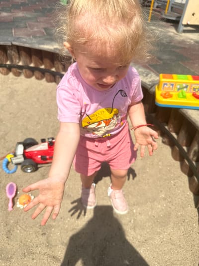 A young child with blonde hair, wearing a pink t-shirt and pink shorts, stands in a sandy area. The child's hands are outstretched and covered in sand. To the left of the child, a red and white toy race car with black wheels and a blue ring is visible. Near the car, a small pink toy brush and a yellow toy with red and green buttons are present. A bright yellow and red plastic toy with various colorful buttons and images is situated to the right of the child, partially obscured by the child's arm. The ground is composed of sand and small pebbles. In the background, a wooden fence-like structure borders the sandy area, with a hint of a playground structure visible further in the distance. The lighting suggests an outdoor, sunny environment. The scene appears to be a playground or sandbox area in Donchovci, Bulgaria.