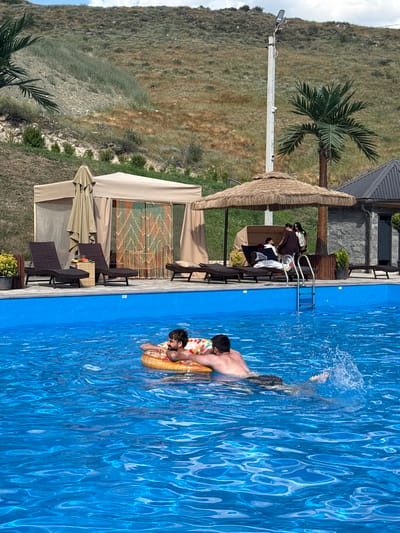This image depicts a sunny outdoor scene at a swimming pool, likely in Verin Ptghni, Armenia, as indicated by the background landscape. Two men are in the bright blue water. One man is lying on a donut-shaped inflatable, his head and arms resting on it, while looking to his right. Another man, positioned behind him and slightly to his left, is swimming and splashing water, his back to the viewer.
In the background, a paved area surrounds the pool. Several lounge chairs are placed on this paved area, with two in the foreground unoccupied. Behind the chairs is a light-colored tent structure with open sides and a beige fabric canopy. A decorative beaded curtain hangs in the entrance of the tent. Further in the background, there's a thatched-roof umbrella covering more lounge chairs where three people are seated. A stone building with a dark roof and windows is visible to the right of the thatched umbrella. A tall lamppost with security cameras is positioned behind the tent and umbrella. The terrain behind the pool area is a grassy, rocky hillside. A palm tree is visible to the right of the lamppost.