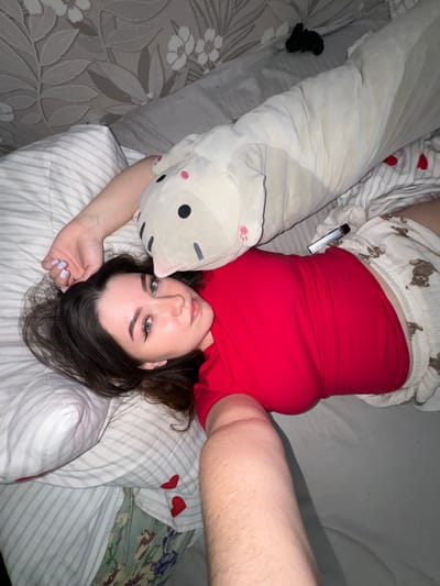 A young woman is depicted lying on her back on a bed, looking directly at the camera. She has dark hair, light-colored eyes, and well-defined eyebrows. She wears a vibrant red sleeveless top and white pajama shorts adorned with small brown bear patterns. Her fingernails are painted a pale blue.
Beside her head is a large, light-colored body pillow shaped like a cartoon cat or animal, featuring black circular eyes and pink ear details. A metallic silver vape pen is visible, tucked into the waistband of her shorts. The bed is covered with an assortment of bedding, including a grey and white floral patterned sheet to her left, a striped sheet beneath her, and a green floral sheet with botanical patterns on the right. Another striped pillow with red heart decals is visible behind her. A small, crumpled black item, possibly clothing, lies on the patterned sheet to the left. The scene suggests a relaxed moment in a bedroom setting, likely captured as a casual selfie or portrait. The location context for this image is Башкултаево, Russia.