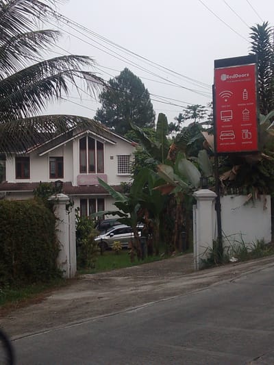 The image shows a white two-story house with a red roof, identified as RedDoorz in Megamendung, Indonesia. A prominent red sign displaying the RedDoorz logo and amenities such as free WiFi, mineral water, television, bathroom, spotless linen, and amenities is visible. The house is surrounded by lush greenery, including palm trees and banana plants. A paved driveway leads to the entrance, where two cars are parked. The sky is overcast, suggesting a cloudy day. The overall scene depicts a tranquil setting in a residential area, likely a hotel or guesthouse.