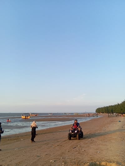 The scene depicts a coastal area identified as a beach in the city of Tritunggal, Indonesia, under a clear, light blue sky consistent with late afternoon or early evening illumination. In the foreground, a sandy beach occupies the lower half of the frame. An adult male operates a black four-wheel All-Terrain Vehicle (ATV) centrally positioned, with a young child seated in front of him. Both individuals face towards the left of the frame. To their immediate left, a person attired in dark clothing and a light-colored hijab stands on the exposed wet sand, facing the body of water. A partially visible individual stands further left. The mid-ground transitions into an expanse of shallow water and exposed mudflats, where multiple traditional fishing boats are moored or beached. These boats vary in color, with at least one prominent yellow-hulled vessel featuring a canopy. Several indistinct human figures are visible wading in the distant shallow waters. The background along the right flank of the beach is characterized by a dense line of coniferous trees. Scattered along the treeline are several makeshift structures, possibly temporary shelters or fishing equipment, and small groups of individuals.
