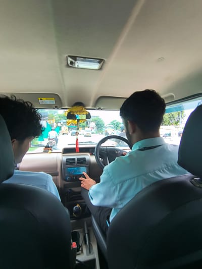 Two men are inside a vehicle, viewed from the back seats. The driver, on the right, is a man with dark hair wearing a light blue collared shirt. He is looking forward and operating the vehicle's infotainment system with his right hand. The passenger on the left has dark, curly hair and is also wearing a light blue collared shirt. He is looking towards the right. The interior of the vehicle is visible, including the dashboard with a touchscreen display, gear shifter, and the car's ceiling with a light fixture. A yellow decorative item hangs from the rearview mirror. Through the windshield, a street scene is visible, suggesting the vehicle is in motion. The street shows other vehicles, including a motorcycle with a rider, buildings, and trees. A "WARNING" sticker is visible on the sun visor. The image appears to be taken in Sidhpur, India, based on the provided context, and depicts a typical urban street scene from within a car.