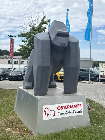 The image shows a large, gray, polygonal sculpture of a gorilla standing on a metallic pedestal. A sign on the pedestal reads "OSTERMAIER Ihre Auto-Familie" with a red outline of a gorilla. Behind the sculpture, a car dealership lot is visible with several cars, including a black Volkswagen ID.3, a yellow car, and a dark gray SUV. In the background, there are buildings, trees, and a cloudy sky. A tall red sign with the text "ostermaier.de" is visible to the left of the sculpture. Blue flags with text are flying in the background. The scene is located in Vilsbiburg, Germany, at the Ostermaier car dealership.