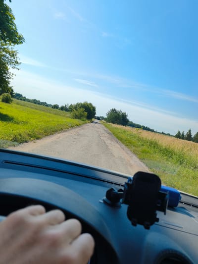 A first-person perspective shot shows a person's hand on a steering wheel, driving down a paved road through a rural landscape. The road is flanked by vibrant green grass and fields of dry, golden grass, with scattered trees and bushes. The sky is a clear, bright blue with a few wispy clouds. The dashboard of the car is visible in the foreground, with a black phone mount and a blue object, possibly a speaker, on the passenger side. The scene appears to be set in the vicinity of Verbuškės, Lithuania, during daylight hours.