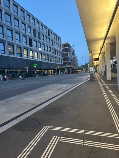The image depicts a modern urban street scene in Liestal, Switzerland, likely at dusk or early evening, given the clear blue sky and prominent artificial lighting. The perspective is from underneath a large, illuminated concrete overhead structure, possibly an elevated walkway or a canopy, which casts a warm glow on its underside.
To the left, a tall, multi-story building with a distinctive, dark grey patterned facade and numerous rectangular windows dominates the scene. Some windows reflect the environment, while others show interior lights. At ground level of this building, several shops are visible. Text seen on the building includes "baloise" and "VISTA" on some windows, and shop signs like "BLKB" and "BAHNHOF APOTHEKE," which features a green cross logo.
To the right, an asphalt road features precise white markings, including sets of triple parallel lines and solid and dashed white lines, suggesting designated lanes or pedestrian areas. Two circular manhole covers are embedded in the road. Adjacent to the road is a concrete sidewalk or platform.
In the mid-ground, a tall, grey information pillar or signpost displays a large number "1" and text, partially legible as "Liestal" and possibly "Bahnhof" or "Busbahnhof." A digital display sign overhead shows "Bus nach Basel SBB," indicating a bus route. A yellow bus can be seen further down the street, and distant hills and buildings are visible against the twilight sky.
Several people are present, engaged in typical urban activities; some are sitting on benches near the shops, while others are standing or walking, suggesting a public transit stop or a busy commercial area. The overall impression is one of clean, well-maintained modern infrastructure.