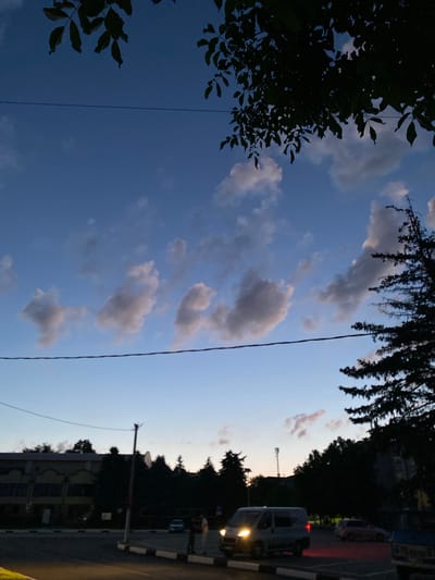 The image depicts an evening scene in Nisporeni, Moldova, characterized by a twilight sky with scattered clouds and silhouetted trees. In the foreground, a paved road is visible with parking spaces marked by white lines and a curb. A light-colored van with its headlights on is parked in the foreground, facing left, with its brake lights illuminated. To the right of the van, a pink car is visible. Two individuals are standing near the parking area, appearing to be conversing. In the background, a multi-story building with a light-colored facade and dark windows is partially visible, along with a dense line of silhouetted trees. A utility pole with wires extends upwards. The sky transitions from a lighter hue near the horizon to a deeper blue overhead, with wispy clouds catching the last light. Dark green foliage from trees hangs over the top of the frame, obscuring part of the sky.