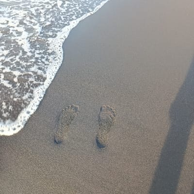 The image shows a sandy beach in Arjasa, Indonesia (latitude: -7.71, longitude: 114.117). The beach is made up of dark grey sand and has a retreating wave of white foam at the left edge of the frame. Two sets of footprints, possibly made by a human, are visible in the sand. They are positioned approximately 1-2 feet apart. The footprints are visible because the sand is wet and there is some water on the beach.  The shadow of a person or object is visible on the right side of the frame, making it unclear whether anyone is present.  The sun is shining brightly, casting shadows.  The image was likely taken during the daytime.