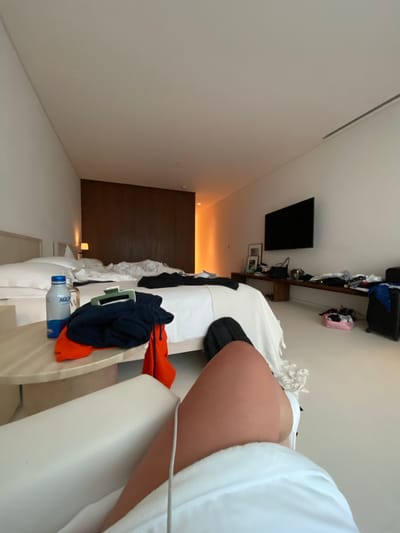 The image shows the interior of a hotel room. A person partially visible in the foreground is sitting in a white chair, wearing a white robe. A bed is visible in the room, covered with white linens and clothes scattered on it. Items such as a water bottle and some clothing are on the bed. A dark-colored television is mounted on the wall. A shelf above the bed is loaded with various items, including suitcases and clothes. A dark wooden paneling feature is present on the wall in the background. The location is unspecified, with no discernible features linking it to El Caracol, Mexico.