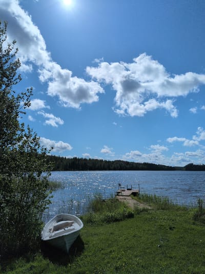 The image displays a serene summer landscape captured in Hartola, Finland. The scene is dominated by a clear blue sky interspersed with scattered white cumulus clouds. The sun, a bright white orb, is prominently visible in the upper left corner, casting a strong glare across the water. A large body of water, likely a lake, occupies the mid-ground and extends towards the background, its surface shimmering with sunlight reflections.
On the left side of the foreground, dense green foliage from trees and bushes partially obstructs the view of the lake. A small, white, wooden rowboat is beached on the grassy shoreline in the lower-left quadrant, resting partly in shadow. The boat's interior shows a single wooden thwart or seat. The foreground consists of lush green grass, with sporadic small yellow flowers.
A wooden pier extends into the lake from the right-center shoreline. A single light-colored chair is positioned on the far end of this pier. The distant shoreline across the lake is lined with a dense forest, primarily composed of dark green coniferous trees. No individuals are present in the image, and no specific actions or interactions are depicted. The environment conveys a tranquil natural setting under bright daylight.