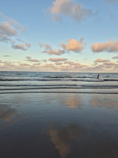 The image depicts a beach scene, likely in Veracruz, Mexico, during either sunrise or sunset. The composition is vertically oriented. The sky is visible on the left, showing a gradient of blue transitioning to a pale yellow-orange horizon, with scattered clouds tinted in shades of peach and grey. The ocean occupies the central portion of the image, featuring gentle waves rolling towards the shore. Several small dark figures are visible in the water, appearing to be people standing or wading. The beach extends across the right side of the frame, its surface reflecting the sky and clouds, creating a mirrored effect. The sand is wet and dark, possibly due to the receding tide. The overall impression is a serene coastal landscape at dawn or dusk.