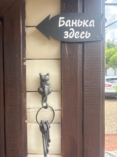 A black arrow-shaped sign is mounted to a wooden post and reads "Баня здесь" (Bathhouse here) in white cursive font, pointing to the left. Below the sign, a cast iron cat figurine is attached to the wall, serving as a hook. A ring with several old keys hangs from the cat's tail. The sign is on a light-colored, horizontal wooden plank wall, with dark brown wooden posts on either side. The background shows greenery, a white fence, and a portion of a white car parked on a gravel driveway. The scene appears to be outdoors, possibly in a rural or suburban setting in Лотос Снт (марьино), Russia.