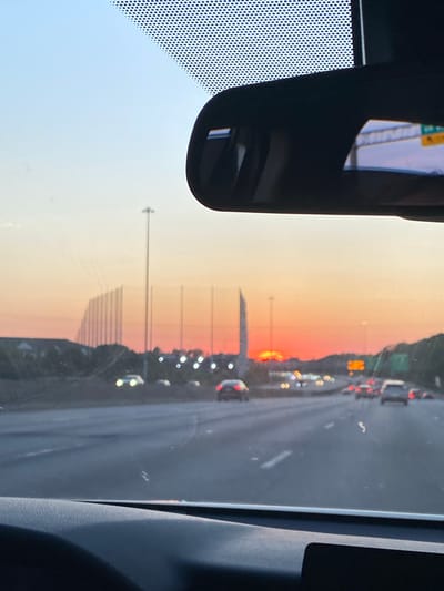 This is a photograph taken from inside a vehicle, looking forward through the windshield. The scene depicts a highway during a sunset. In the foreground, the dashboard of the car is visible. The highway stretches into the distance, with several cars on it, some with their headlights on. On the left side of the highway, there is a fence structure with tall poles and netting, possibly for a driving range or sports facility. A tall, slender monument or building is visible in the middle distance. The sky is a gradient of pale blue to orange and red, indicating the sun is setting. The location is identified as the city of Virginia Beach, United States of America.