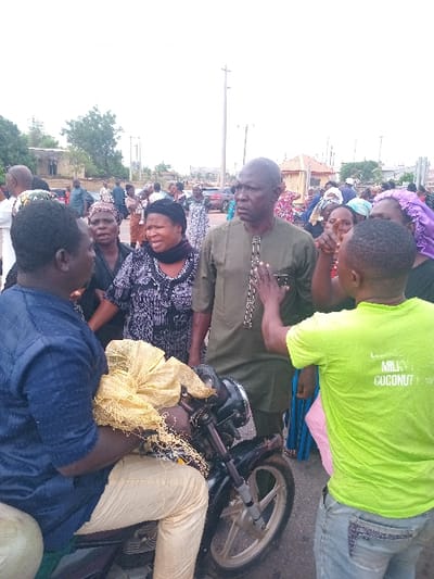 The scene is a street in Ungwan Tanko, Nigeria, a small village in Kaduna State. The coordinates provided, 10.422 latitude and 7.49 longitude, confirm the location. There are nine individuals in the scene, mostly adults, with one person seated on a black motorcycle. The individual on the motorcycle is wearing a blue shirt and khaki pants, with a large yellow bag over his left shoulder. In front of him are two women, one wearing a patterned black and white dress with her hair covered, and the other with a black dress and a scarf tied around her head. These two women are talking to a man wearing a green shirt and blue jeans. He is gesturing with his right hand towards the motorcycle. Beside him, a younger man is wearing a bright green shirt, and is gesturing towards the motorcycle with his right hand. Two other individuals, wearing colorful attire, stand in the background. In the distance, buildings and trees are visible. The street appears to be paved, but some sections are cracked. The weather appears to be overcast, with low-lying clouds and bright sun. There is no rain present.