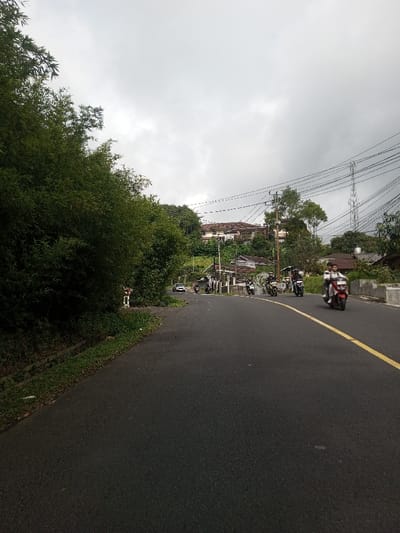 An outdoor scene in Saing Ta Mawt Lay, Myanmar, depicts a paved two-lane road under an overcast sky. The road features a solid yellow line delineating the right side of the visible lane. To the immediate left, dense green foliage, primarily bamboo, borders the roadway above a narrow grassy verge and a concrete drainage channel. On the right, the road runs alongside a series of low concrete barriers. In the mid-ground and background, multiple structures, including residential buildings with various roof styles, are visible on an elevated, tree-covered hillside. A large, multi-story building with a prominent reddish-brown roof stands atop a distant hill. Overhead utility poles with numerous power lines traverse the upper portion of the scene, and a tall metal lattice communication tower is visible in the far right background.
Vehicle traffic is present on the road. In the foreground, a red scooter carries two individuals, traveling away from the viewer in the right lane. Multiple additional motorcycles, some appearing to be scooters and others standard motorcycles, are visible further down the road, with some traveling in the same direction as the red scooter and others potentially approaching. A white sedan is partially visible further down the road on the left side, near a bend.