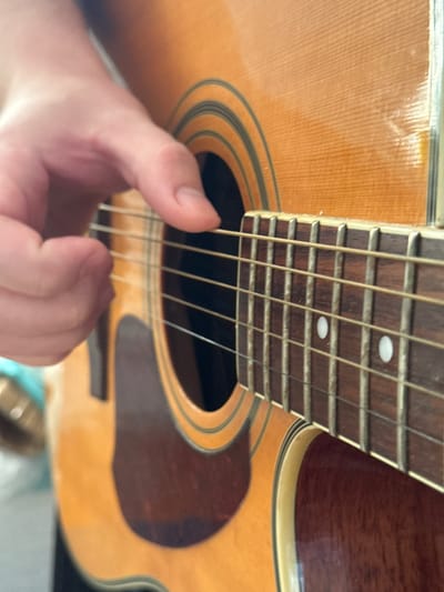 The image shows a close-up of a person's hand playing an acoustic guitar. The focus is on the fretboard and the strings. The guitar has a light brown wooden body with decorative black and white binding around the soundhole. The fretboard is dark wood with frets and two circular white inlays. The hand appears to be pressing down on the strings on the fretboard, suggesting the action of playing music. The background is out of focus but suggests an indoor setting. There is no specific information in the image to indicate the location in Troitske, Ukraine.