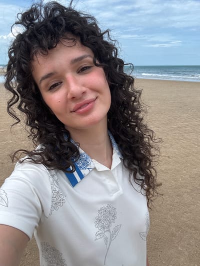 A young woman with long, dark, curly hair is taking a selfie on a beach. She is wearing a white polo shirt with a floral pattern and a blue and white striped collar. The background shows a sandy beach, the ocean, and a cloudy sky. The location is identified as Ban Bang Ket, Thailand.