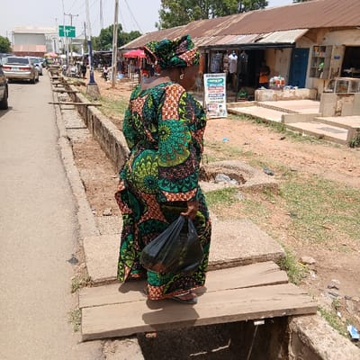 The image shows a woman wearing a green and black patterned dress and a head wrap, walking on a wooden plank bridge over a drainage ditch. She is carrying a black plastic bag in her right hand. The scene is located in a city in Nigeria. The road in the background is paved and leads toward a series of buildings that appear to be shops. The woman is walking away from the camera toward the shops. There are other people and cars visible in the distance. The environment appears to be dusty and somewhat neglected.